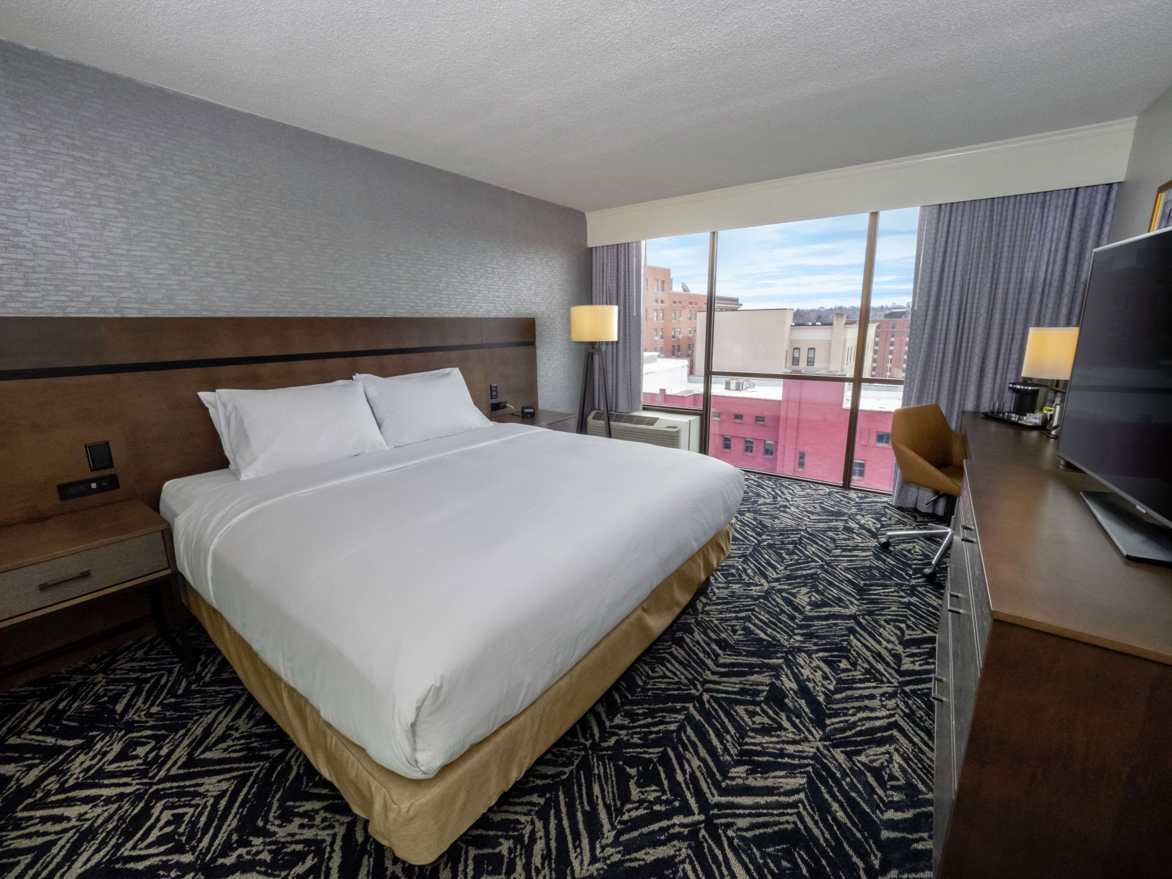 Une chambre d’hôtel avec un grand lit, un bureau avec une télévision, une chaise et une fenêtre donnant sur la ville.