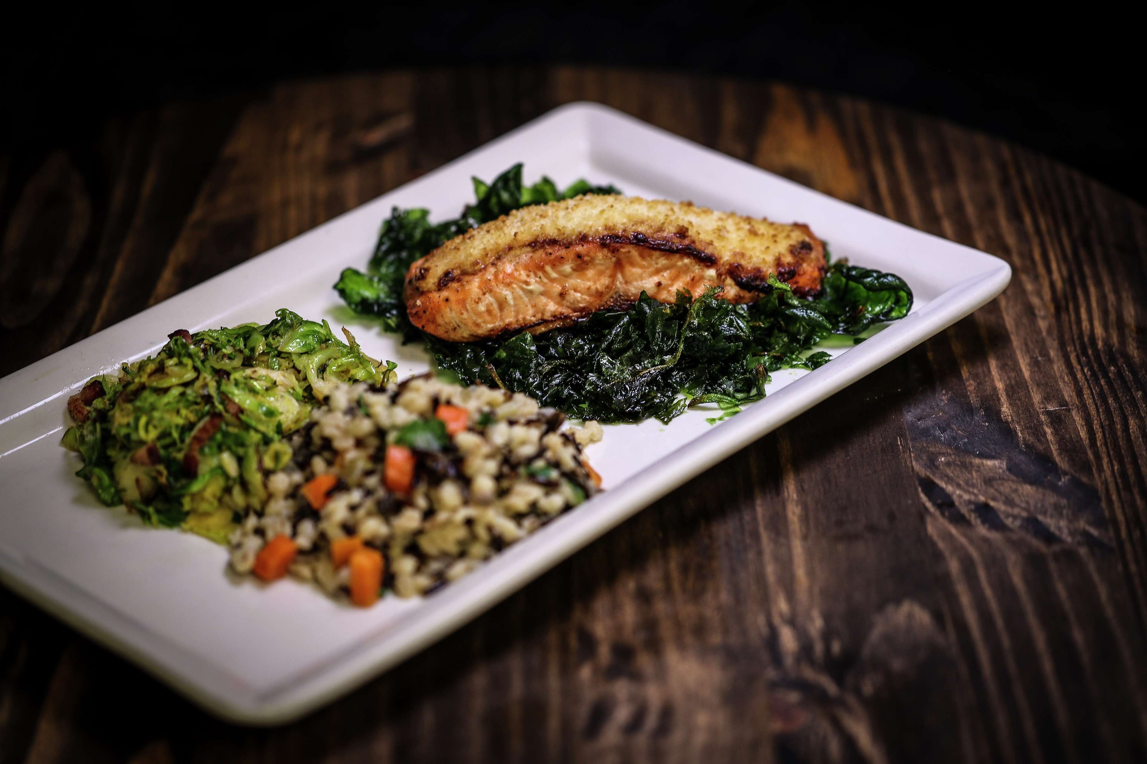 A white rectangular plate with a grilled salmon fillet, a side of sautéed greens, and a grain dish on a dark wooden table.