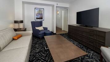 A modern hotel room with a sofa, armchair, coffee table, TV, and a patterned carpet.