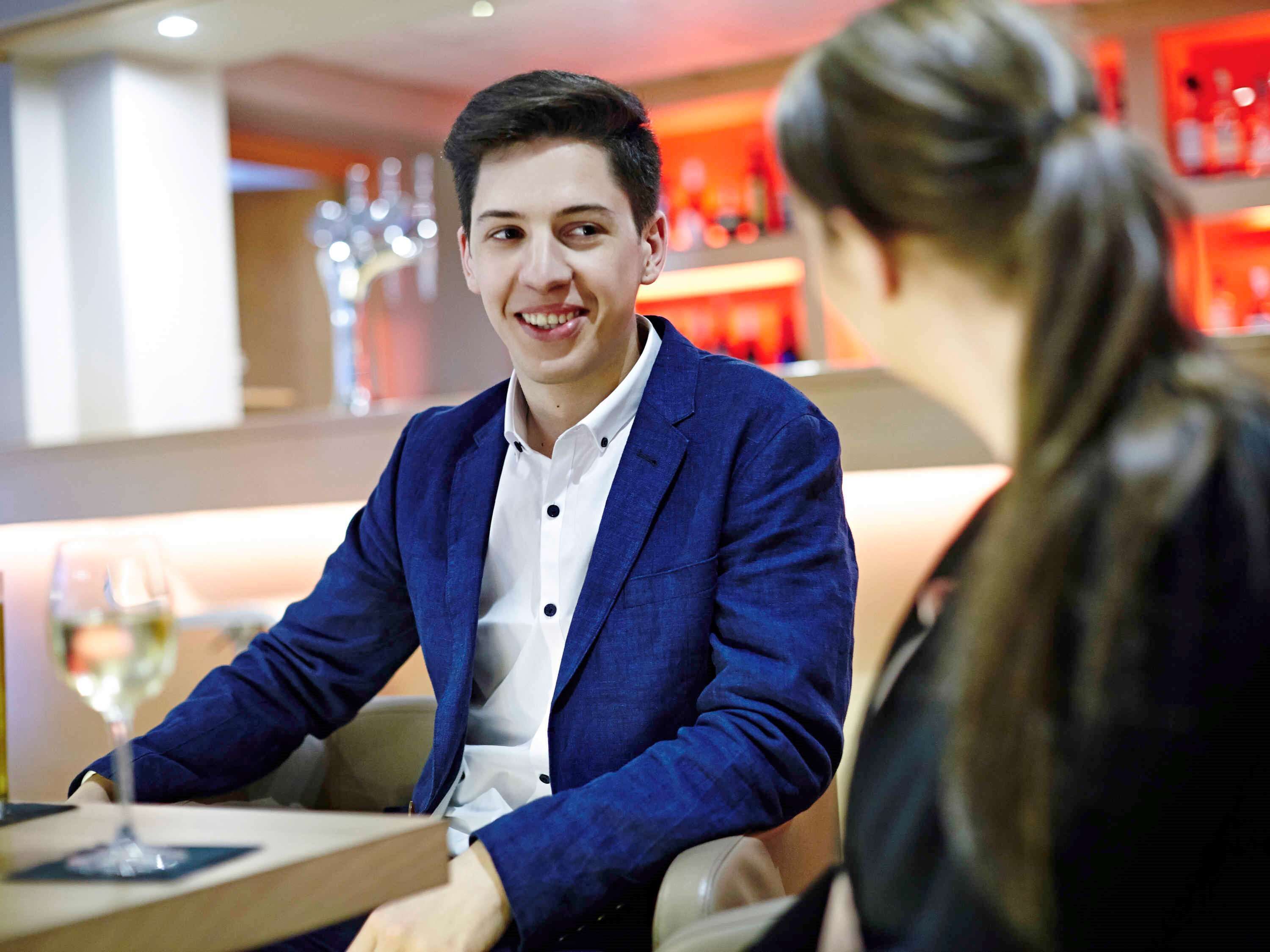 A man in a blue blazer and white shirt sitting at a table with a wine glass, in a room with a red and white color scheme and modern decor.