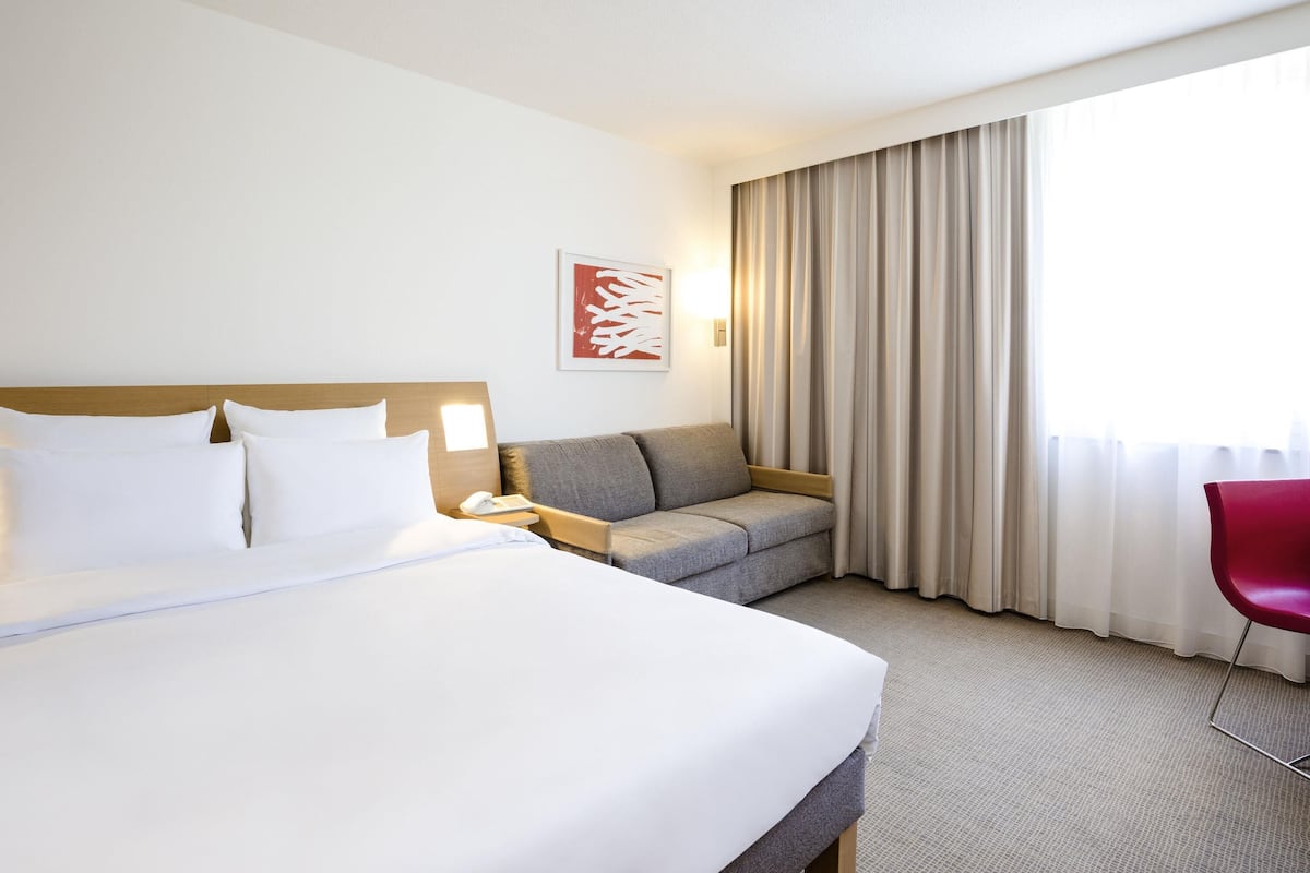 A hotel room with a large bed, a sofa, a chair, a bedside table, a lamp, and a window with curtains.