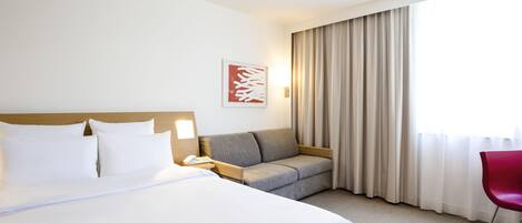 A hotel room with a large bed, a sofa, a chair, a bedside table, a lamp, and a window with curtains.