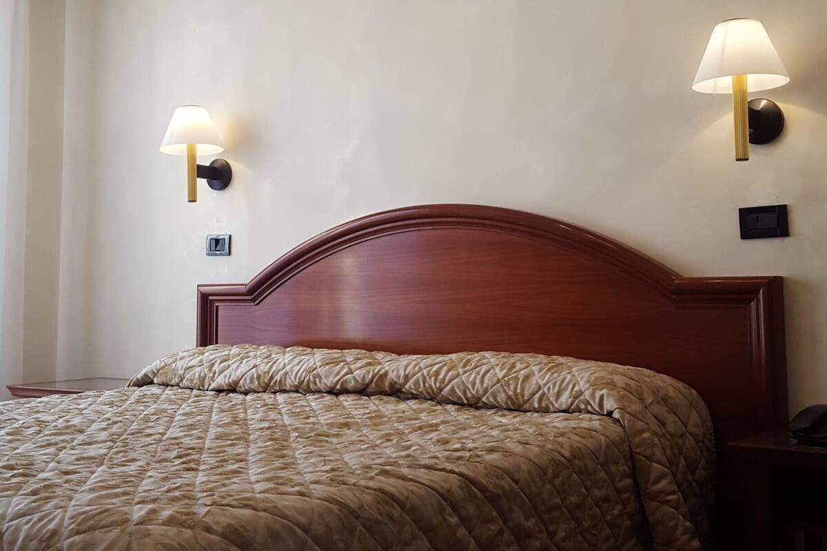 A hotel room with a wooden headboard, a quilted bedspread, a chandelier, and two wall-mounted lamps.