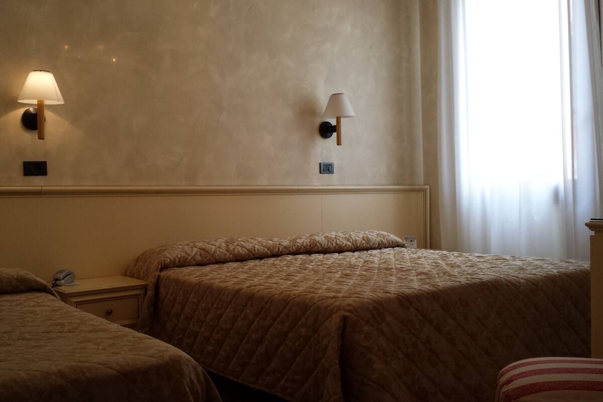 A hotel room with two beds, a nightstand, a lamp, and a window with curtains.