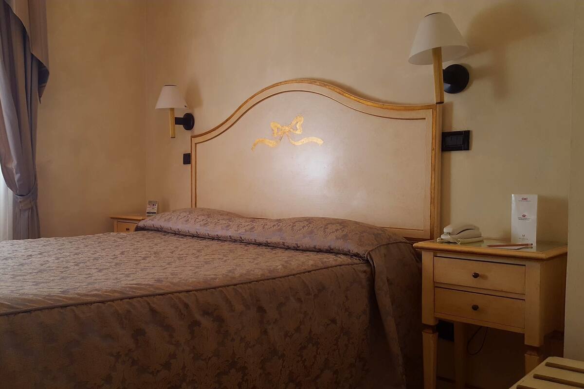 A hotel room with a large bed, two bedside lamps, a nightstand with a phone and a box, and a curtain on the window.