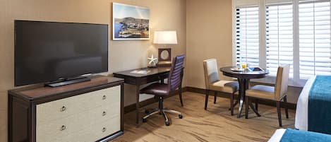 A hotel room with a flat-screen TV, a desk with a chair, a dining table with chairs, and a large window with blinds.