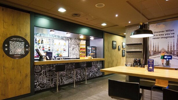 A bar area with a QR code for online orders, a counter with a menu, and a poster advertising Milan. - a&o Hostel Milano Ca Granda (Milan)
