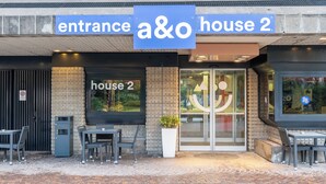 An entrance sign for 'a&o house 2' with a brick facade, glass doors, and outdoor seating area with tables and chairs. - a&o Hostel Milano Ca Granda (Milan)
