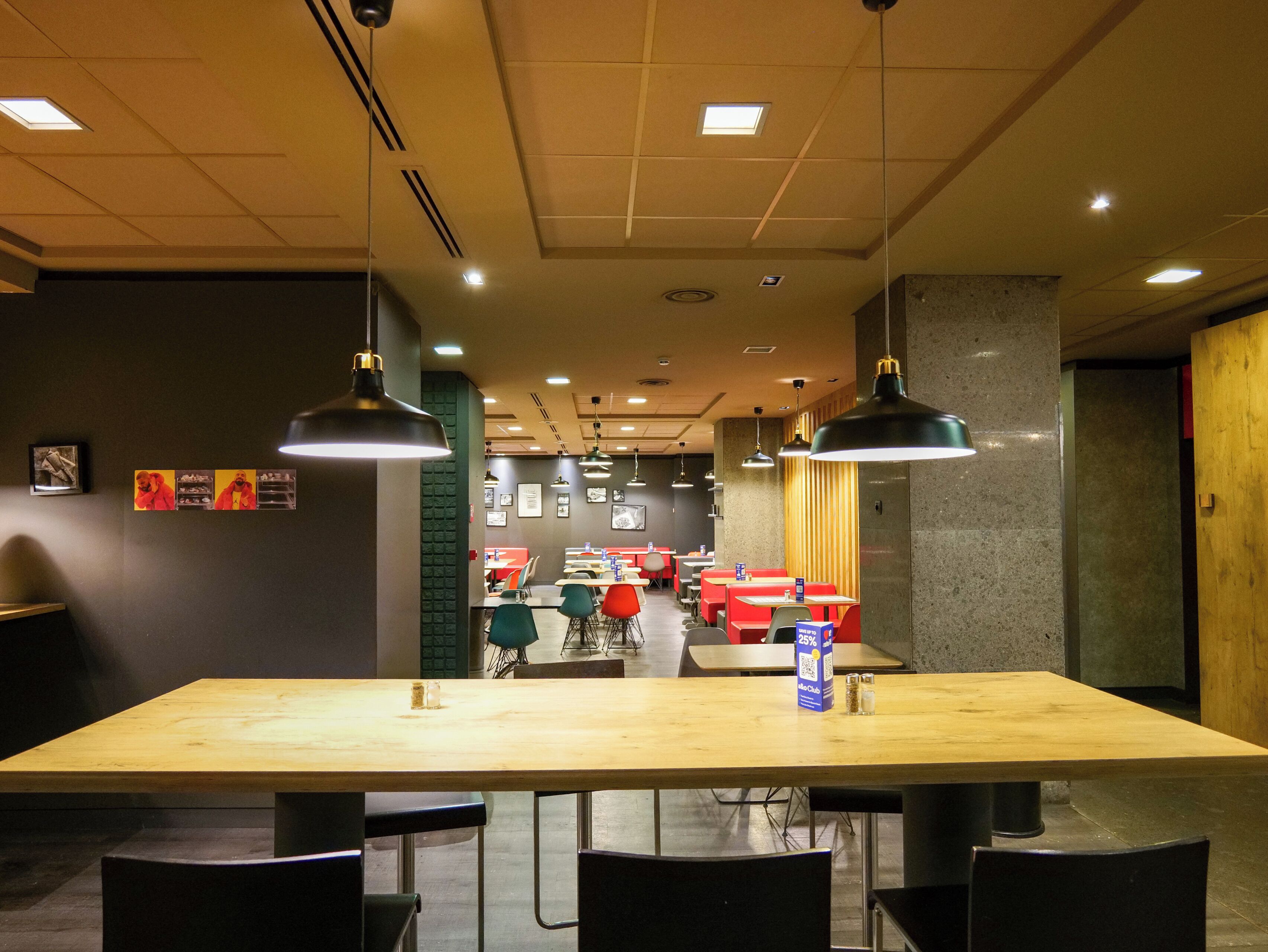 A restaurant interior with a long wooden table, black chairs, hanging pendant lights, and framed pictures on the wall.