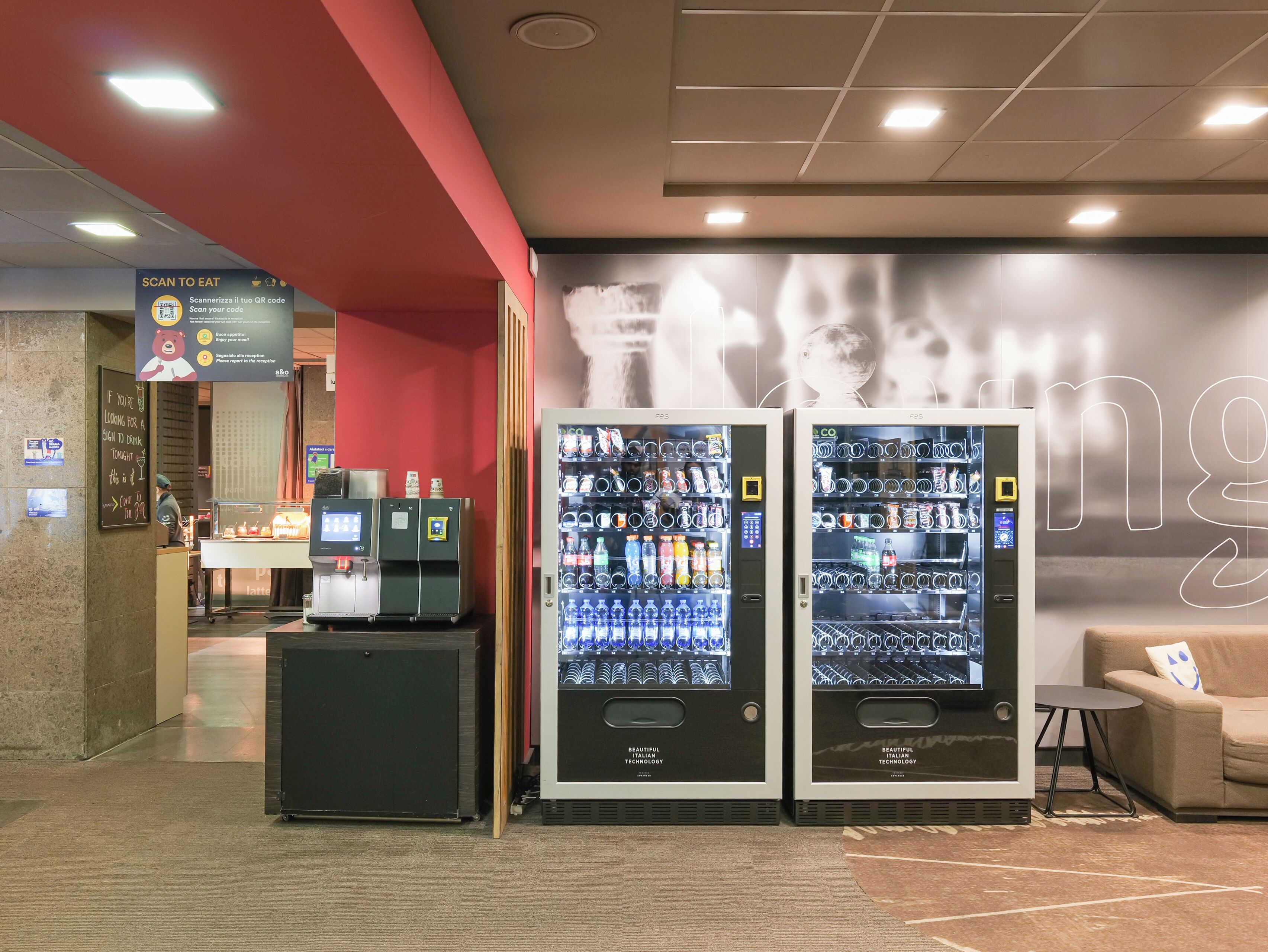 A modern indoor area with vending machines stocked with beverages and snacks, a sign indicating a QR code for scanning, and a reception desk.
