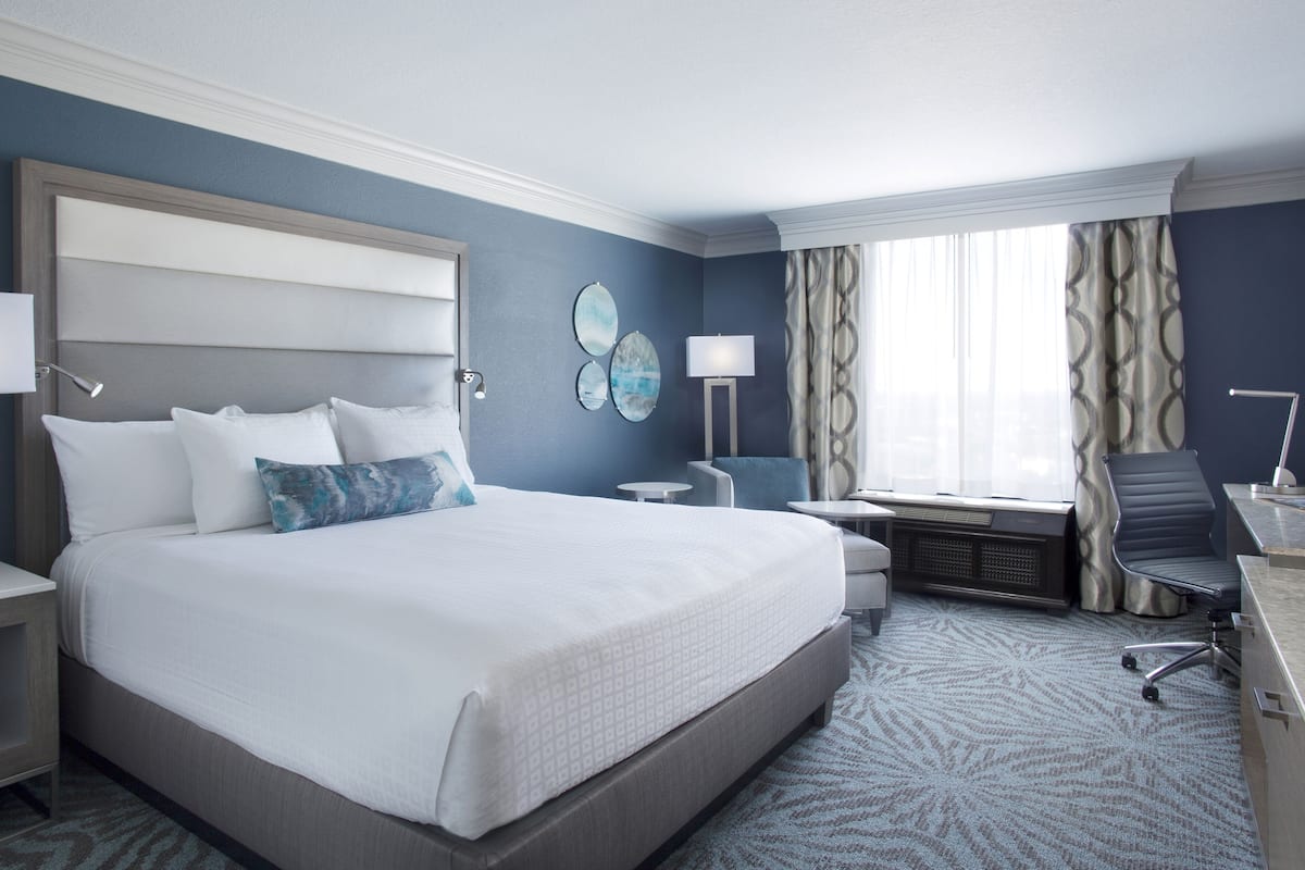 A hotel room with a large bed, bedside tables, a desk, a chair, and a window with curtains.
