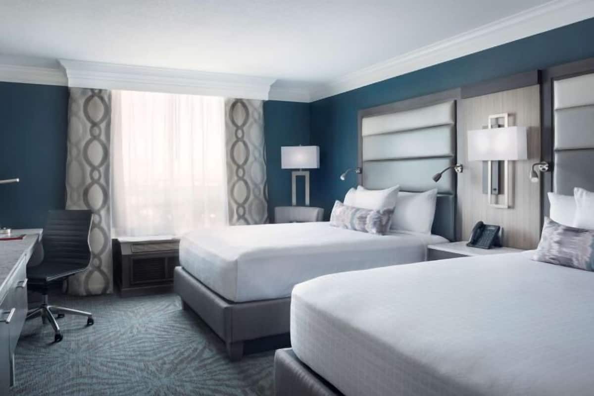 A hotel room with two beds, a desk, a chair, and a lamp.