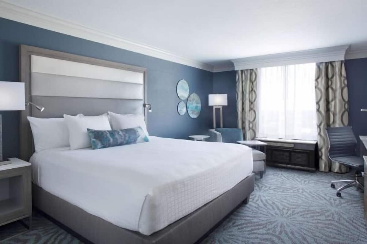 A hotel room with a large bed, bedside tables, a desk with a chair, a window with curtains, and a wall-mounted artwork.