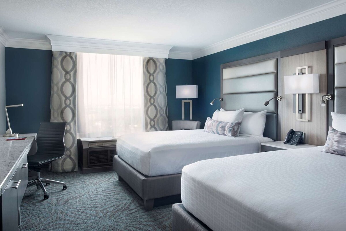 A hotel room with two beds, a desk, a chair, and a lamp.