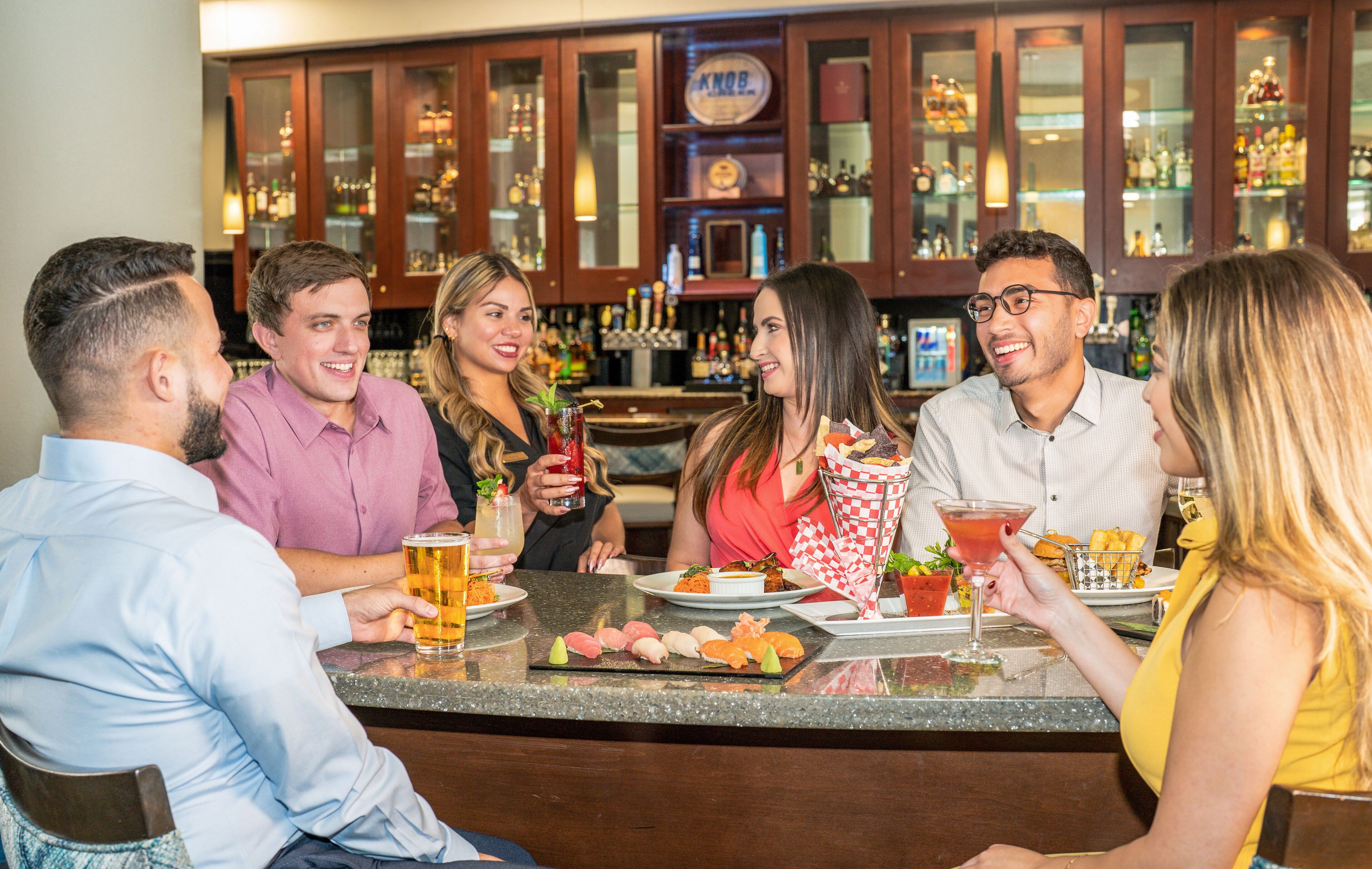 A group of people seated around a bar counter with various drinks and appetizers.
