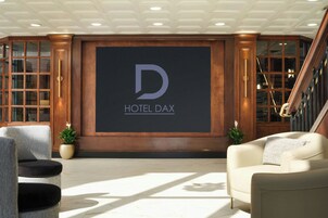 Un vestíbulo con una pantalla grande mostrando el logotipo de HOTEL DAX, una escalera y zonas de estar con plantas en macetas.