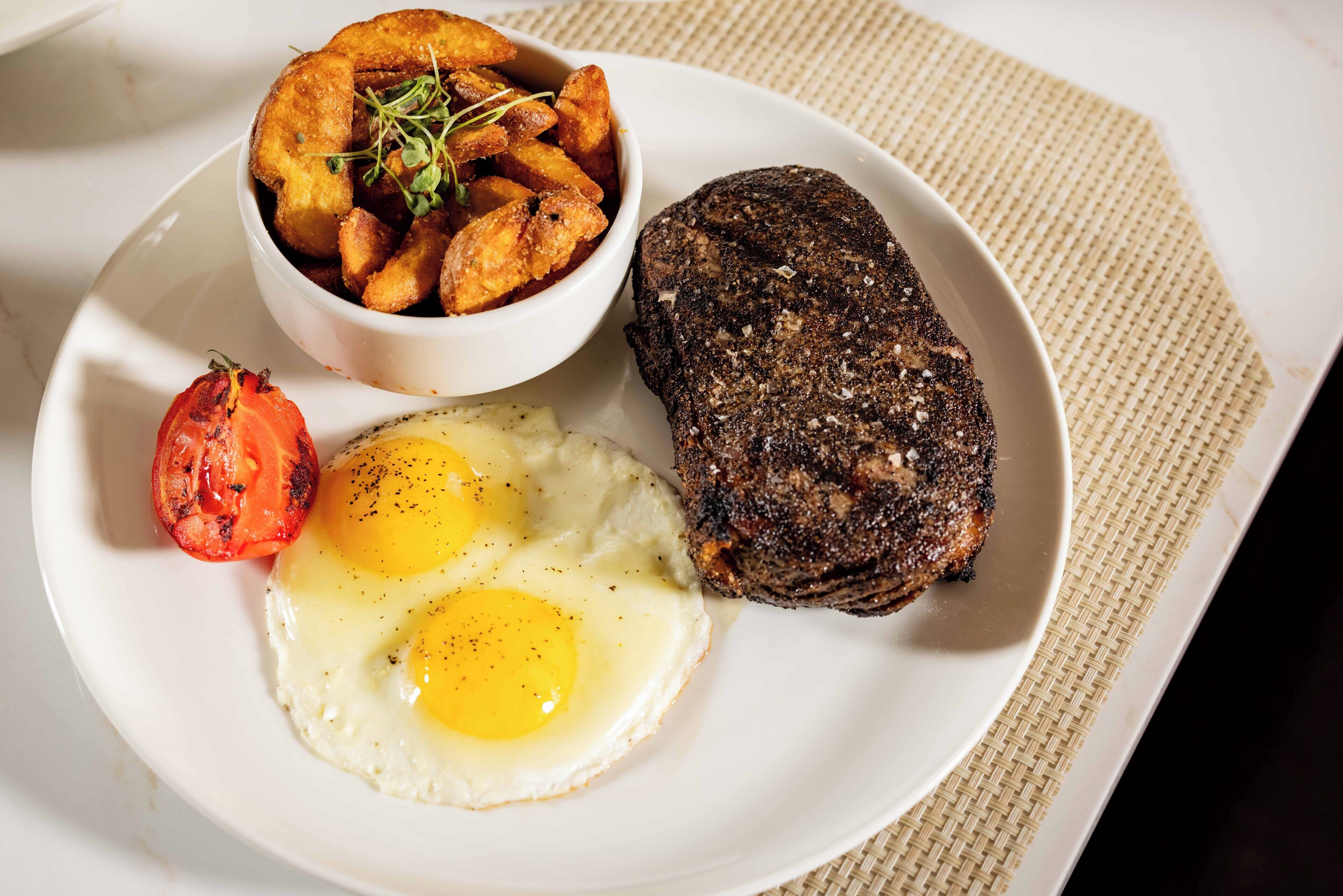 Un plato con un bistec, dos huevos fritos, patatas asadas y un tomate a la parrilla.