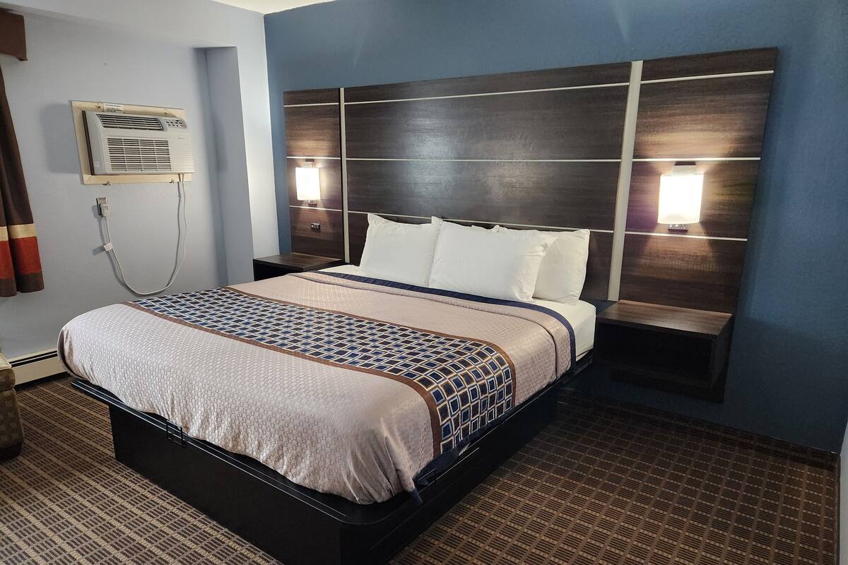 A hotel room with a bed, bedside lamps, an air conditioning unit, and a carpeted floor.
