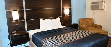 A hotel room with a large bed, two bedside lamps, a nightstand, a chair, and an air conditioning unit.