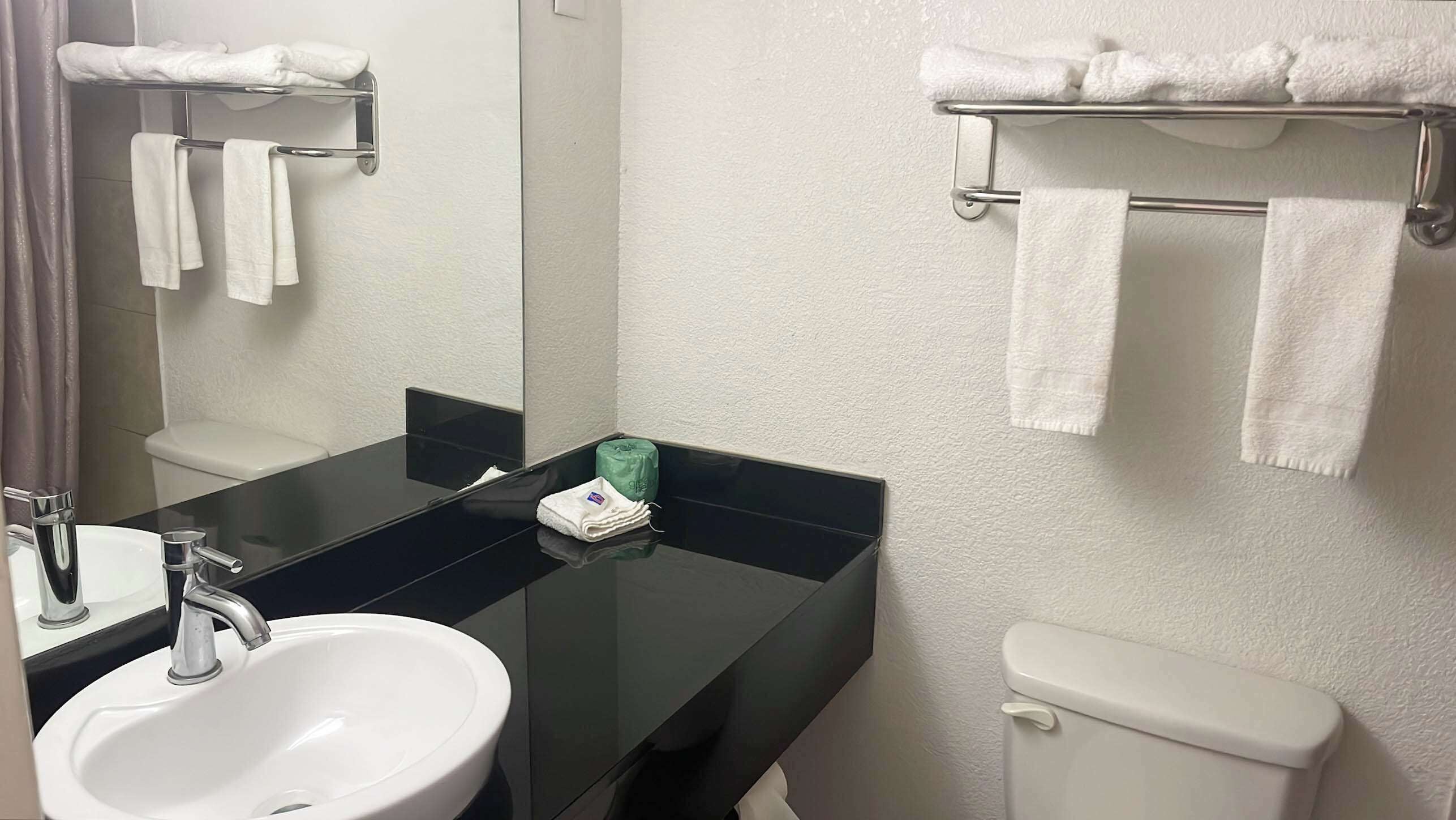 A bathroom with a white sink, a mirror, a black countertop, a toilet, and towel racks with white towels.