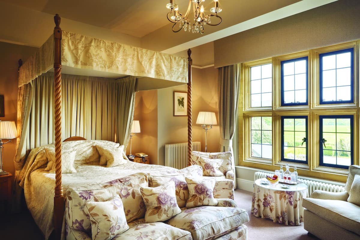 Una cama con dosel y cortinas florales, una mesa redonda con mantel de flores y vistas a zonas verdes a través de amplias ventanas.