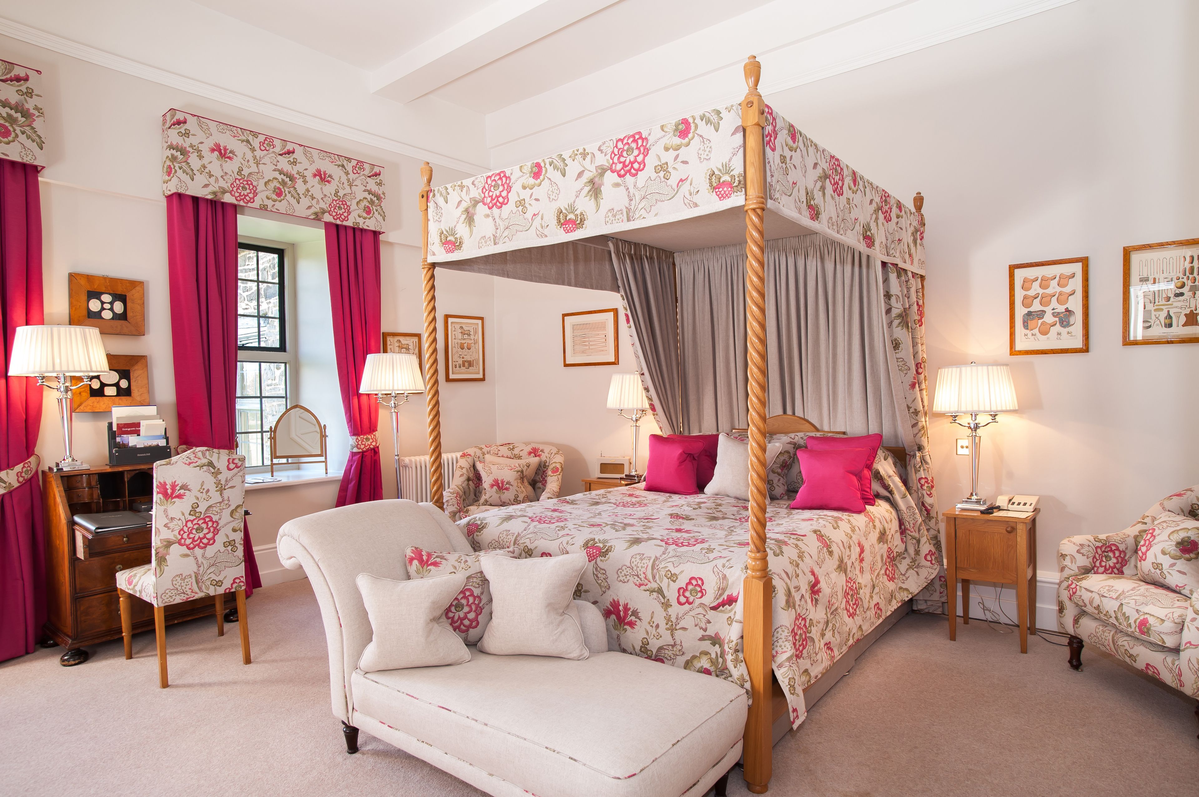 A four-poster bed with floral bedding, a sitting area with a chair and a desk, and artwork on the walls.