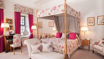 A four-poster bed with floral bedding, a sitting area with a chair and a desk, and artwork on the walls.