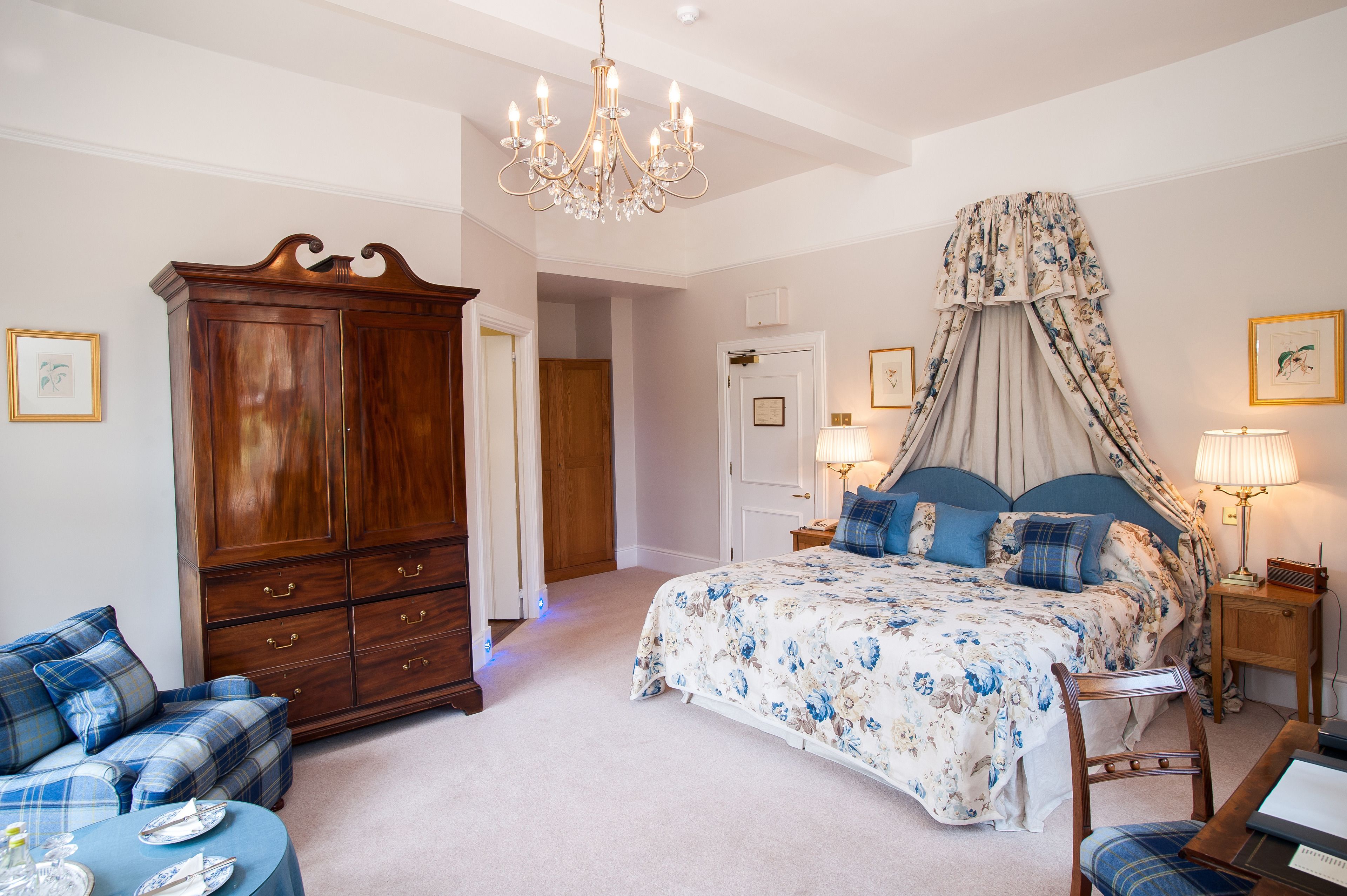 A bedroom with a large wooden wardrobe, a four-poster bed with a canopy, a blue plaid sofa, and a small table with a lamp.
