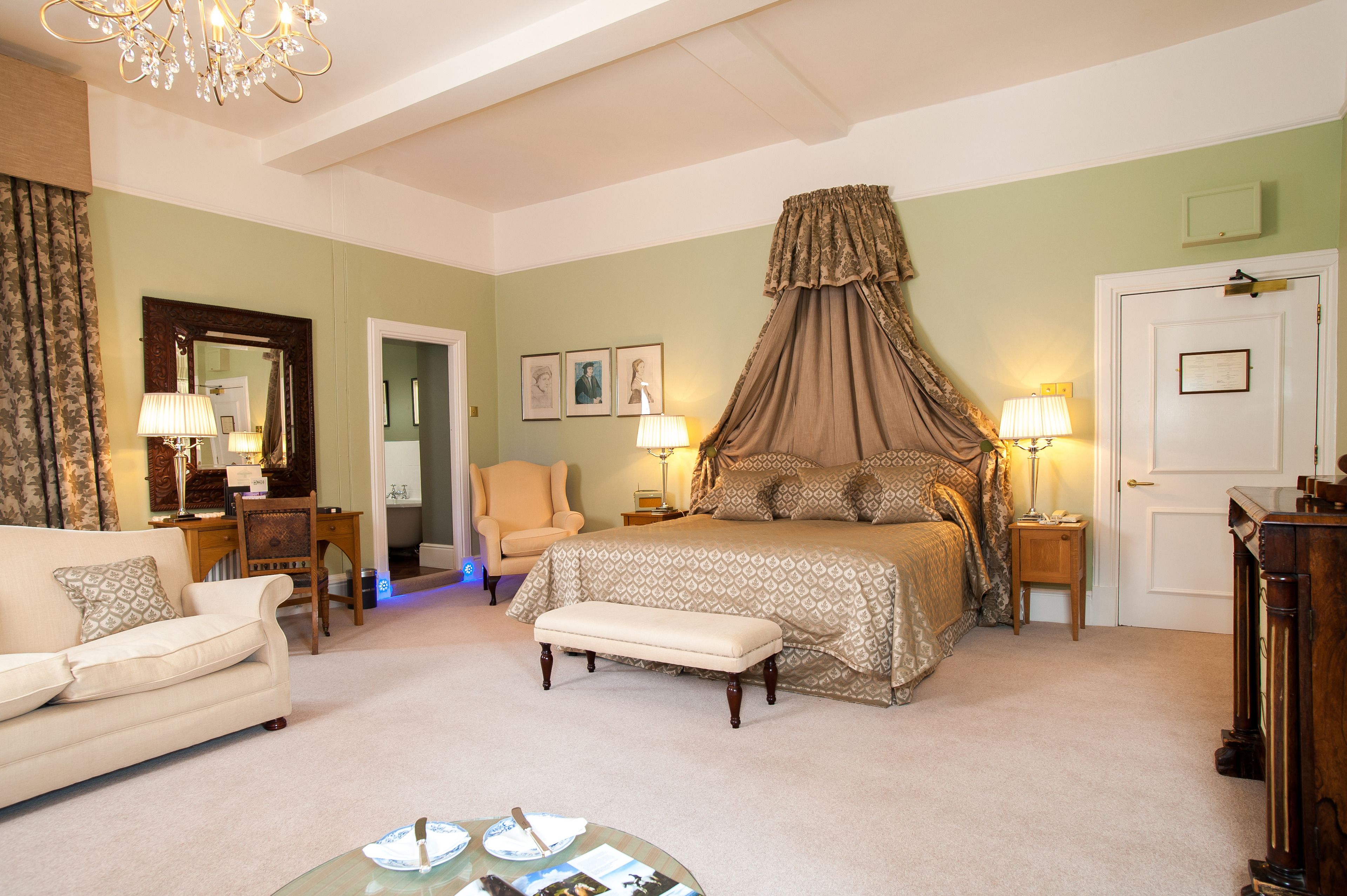 A bedroom with a four-poster bed, a sitting area with a chair and a table, and a door with a mirror and framed pictures.