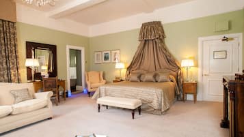A bedroom with a four-poster bed, a sitting area with a chair and a table, and a door with a mirror and framed pictures.
