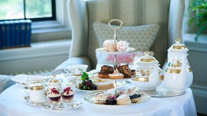 A three-tiered tea service set on a table with various pastries and desserts, a teapot, and a pitcher, in a room with a window and a chair in the background. - Llangoed Hall (Brecon)