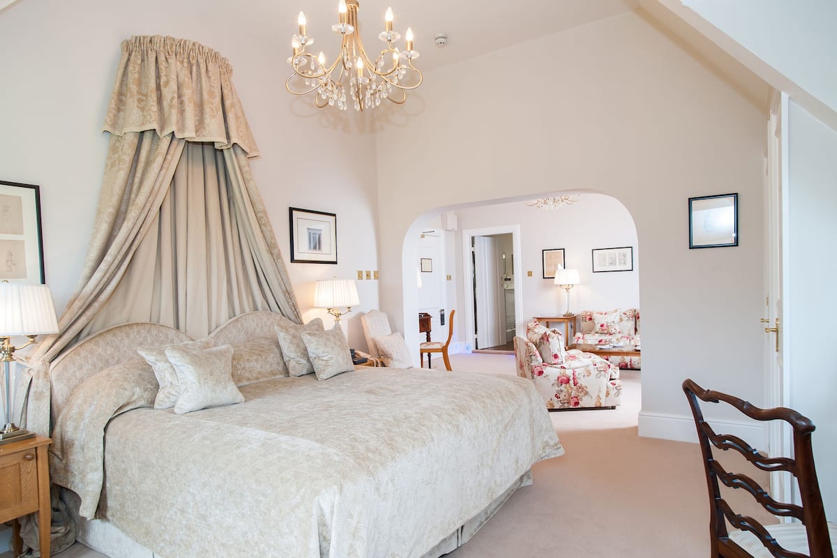 Un dormitorio amplio con una cama grande, una araña y vistas a otra habitación con un sillón floral.