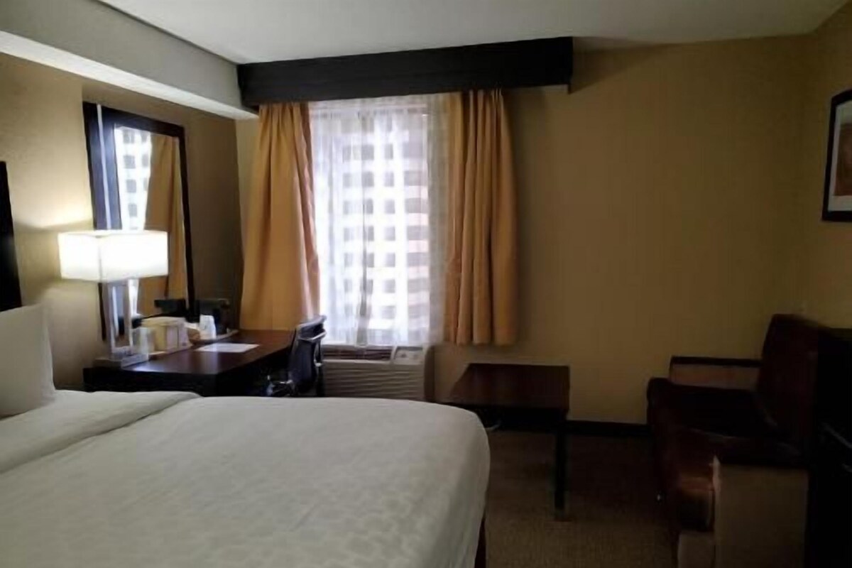 A hotel room with a large bed, a desk with a lamp, a chair, and a small table.