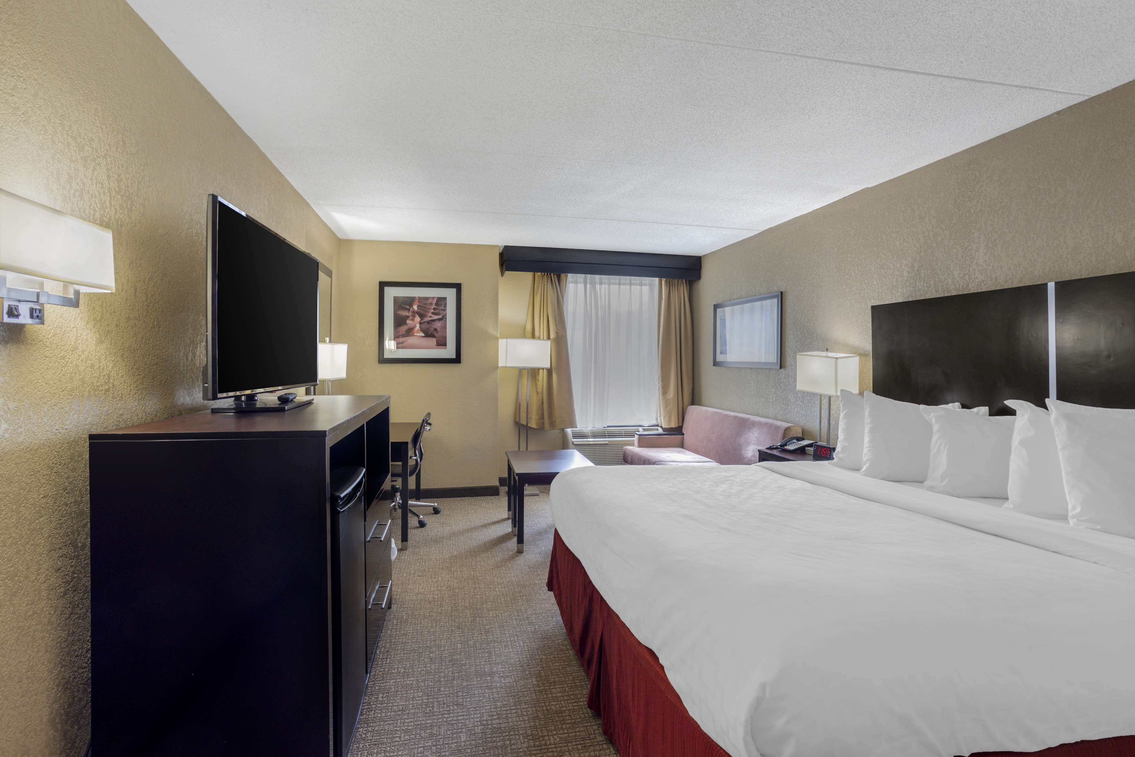 A hotel room with a large bed, a desk with a TV, a chair, a window with curtains, and a flat-screen TV on the wall.
