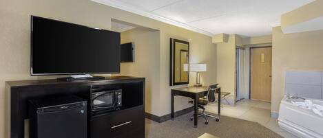 A hotel room with a flat-screen TV, a mini-fridge, a desk with a lamp, a chair, and a bed with a white headboard.