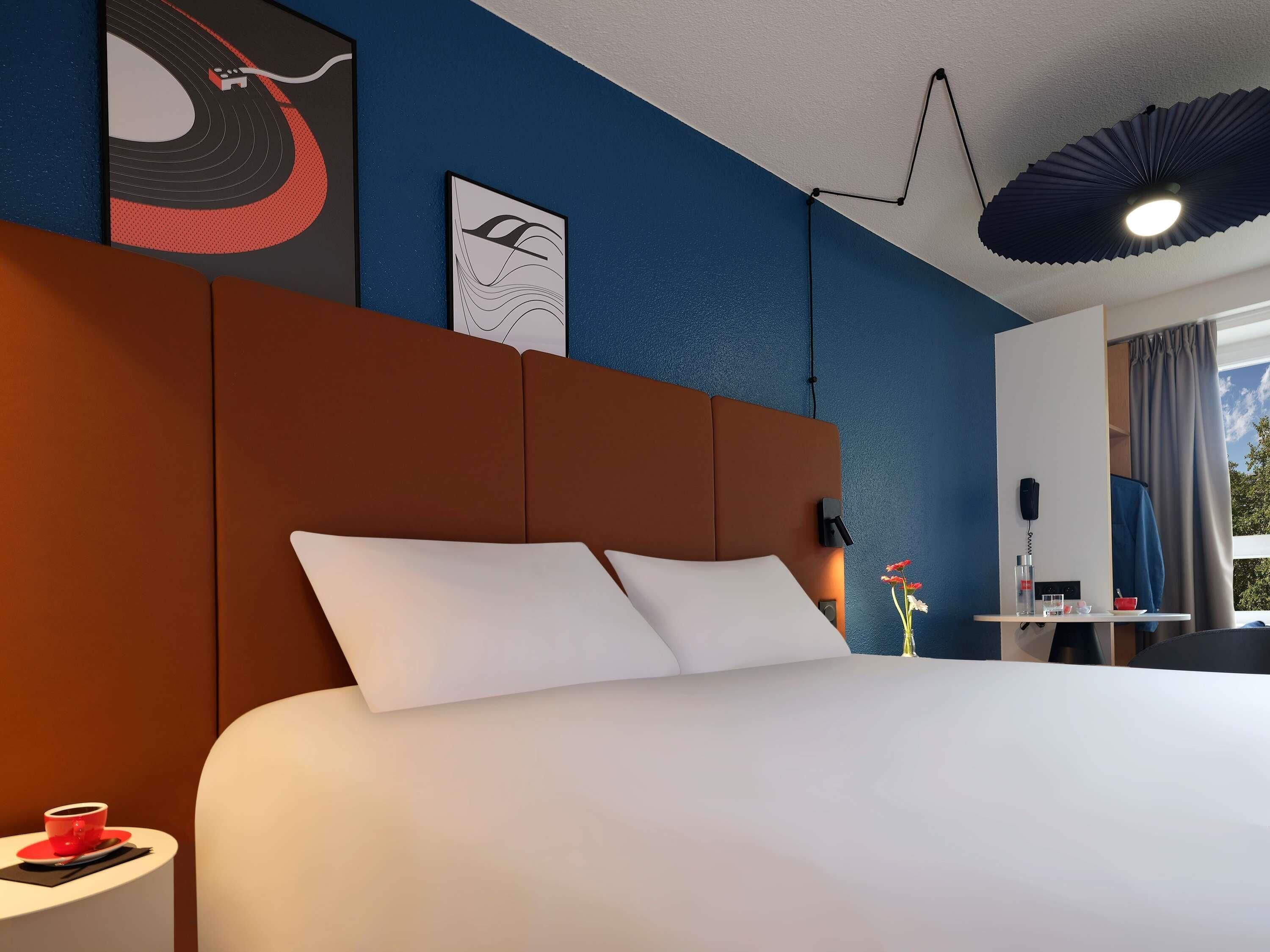 Una camera d'albergo con un letto grande, un comodino con una tazza rossa, un telefono fissato al muro e una ventola a soffitto.