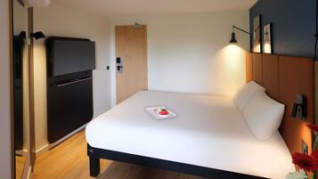 Quarto de hotel moderno com cama, televisão, porta de madeira e uma pequena mesa com uma xícara e flores.