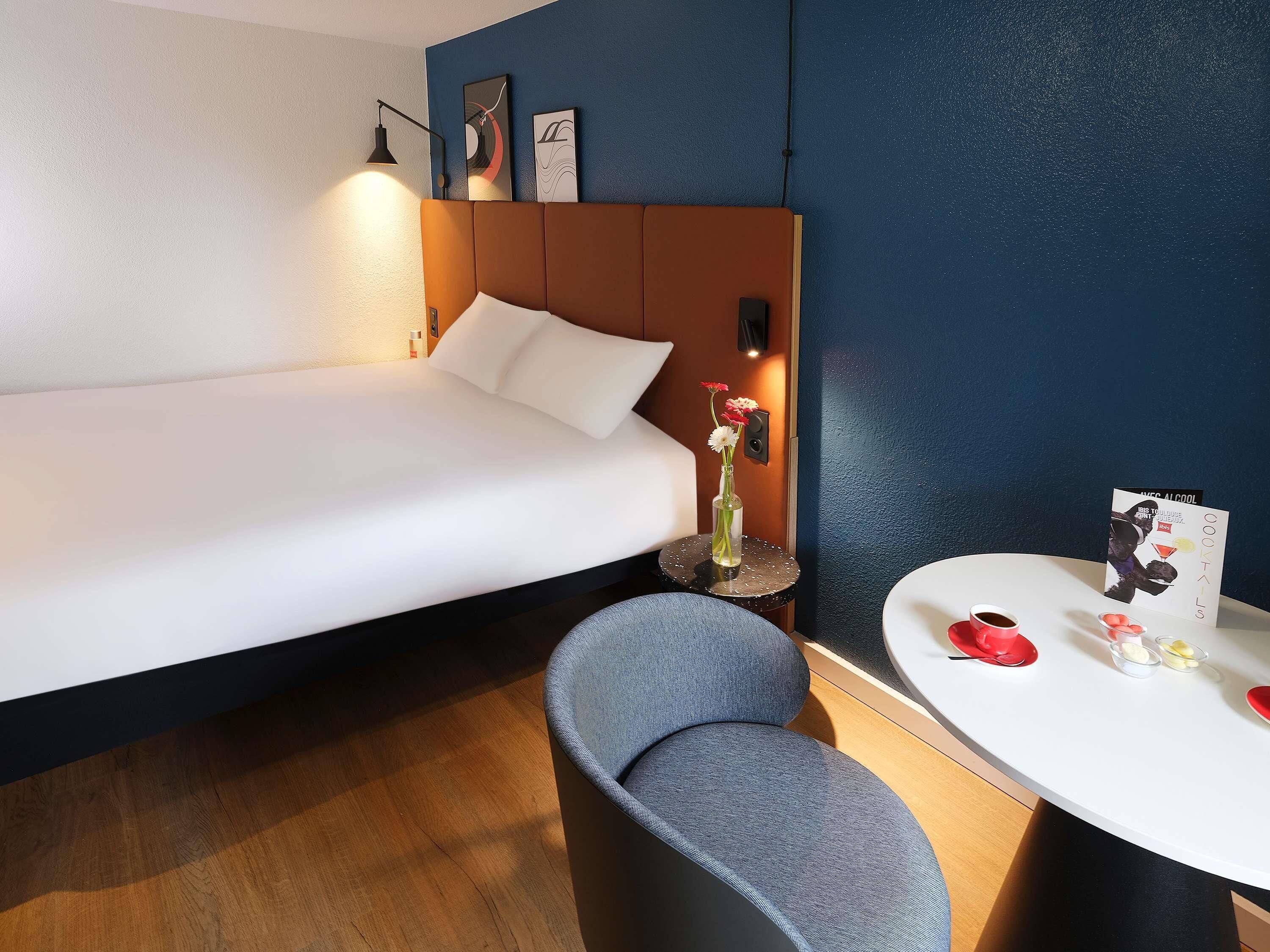 A hotel room with a bed, a chair, a small table with a card and glasses, and a vase with flowers.