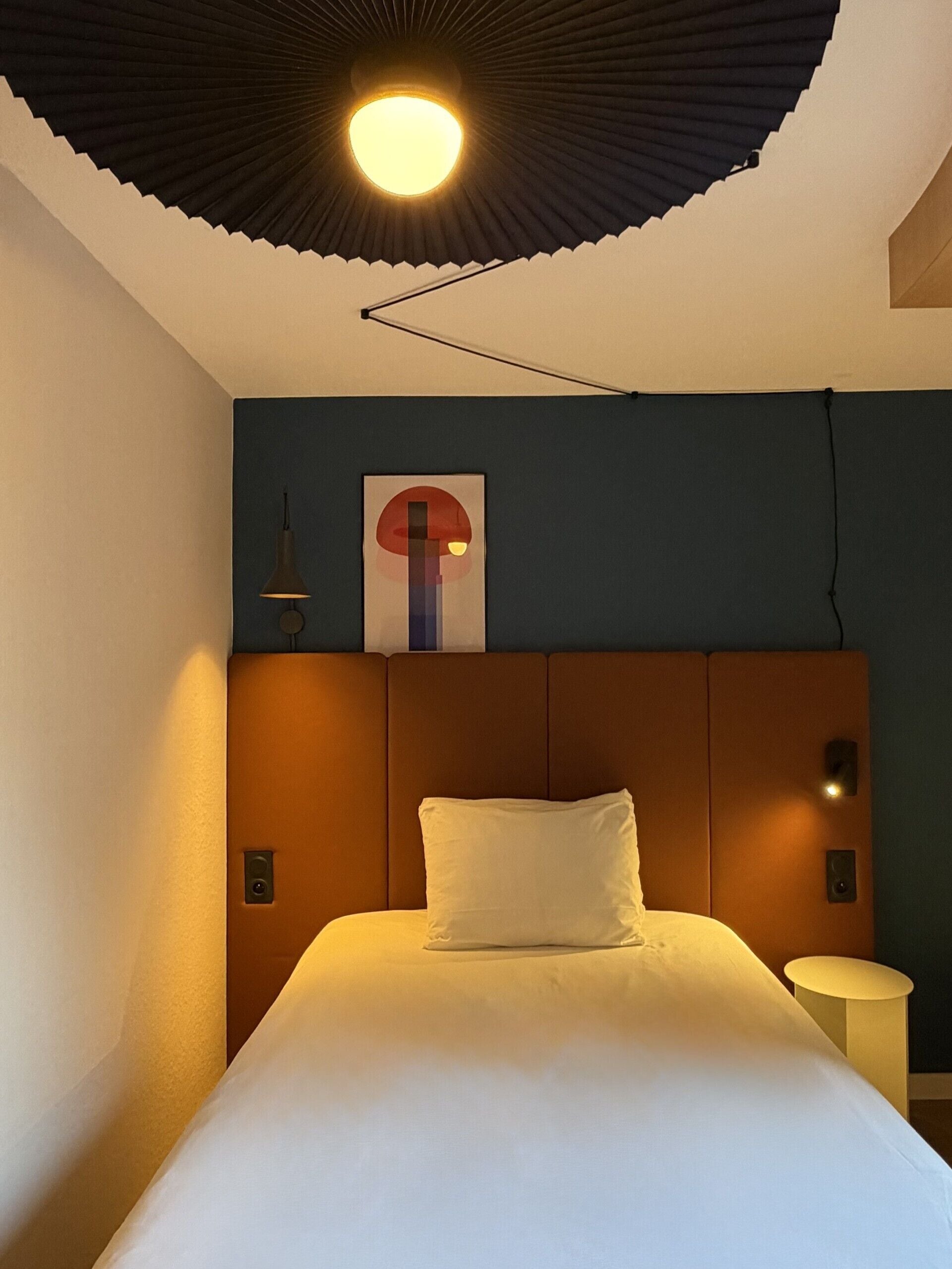 Una habitación de hotel con una cama, una obra de arte en la pared, una lámpara en el techo y una mesita de noche.