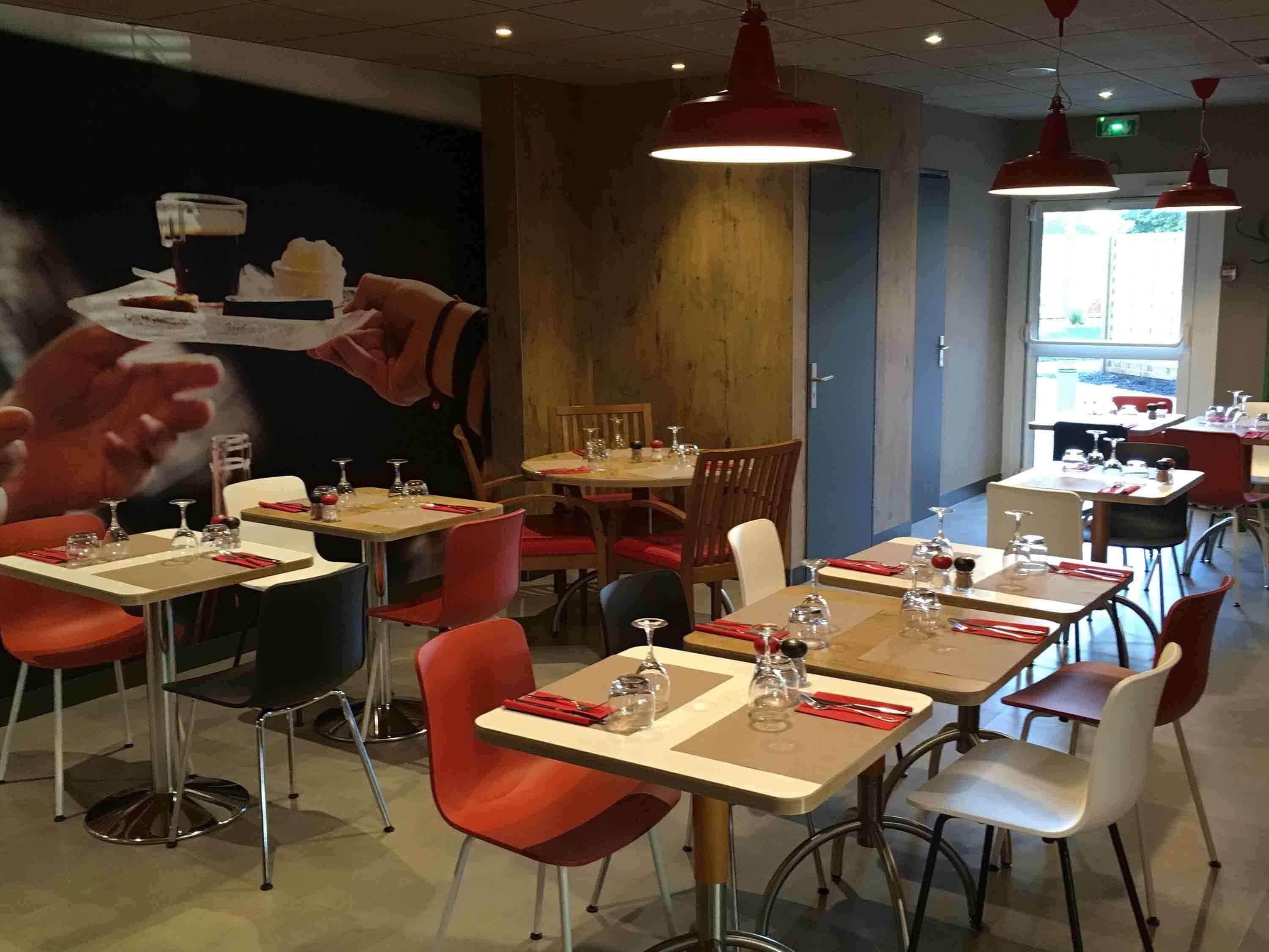 Interior de un restaurante con mesas preparadas para comer, sillas rojas e iluminación colgante.