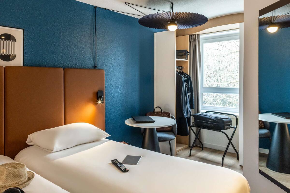 Ett hotellrum med en säng, ett nattduksbord och ett litet skrivbord med en stol. Det finns ett fönster med gardiner, en klädhängare och en resväska.