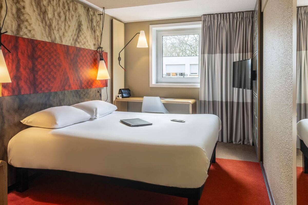 Ett hotellrum med en säng, ett skrivbord, en stol, en tv och ett fönster med gardiner.