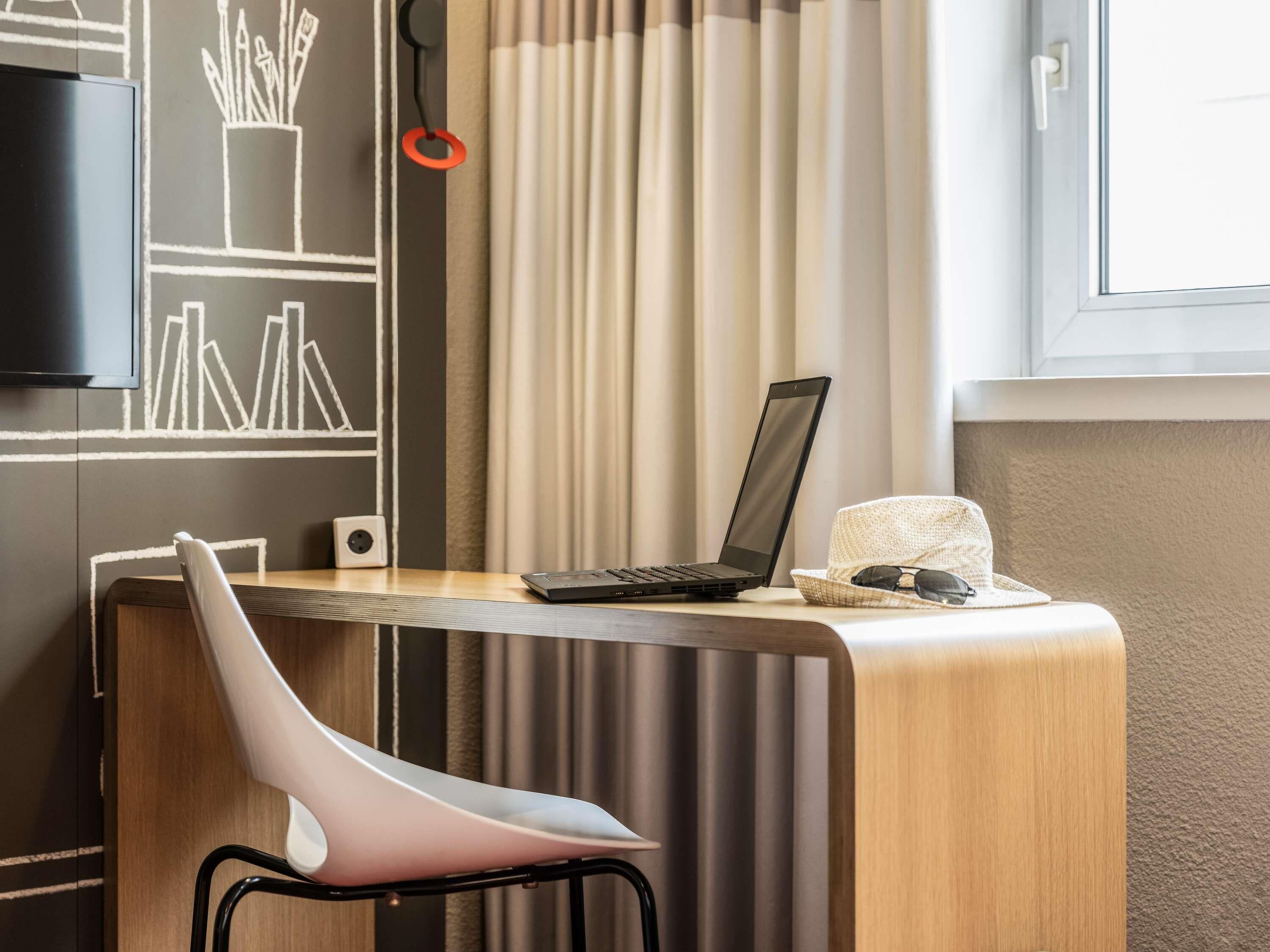 Ein modernes Zimmer mit Schreibtisch, Laptop, Stuhl, Fernseher und einem fensterbepflanzten Fenster.