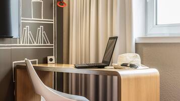 Ein modernes Zimmer mit Schreibtisch, Laptop, Stuhl, Fernseher und einem fensterbepflanzten Fenster.