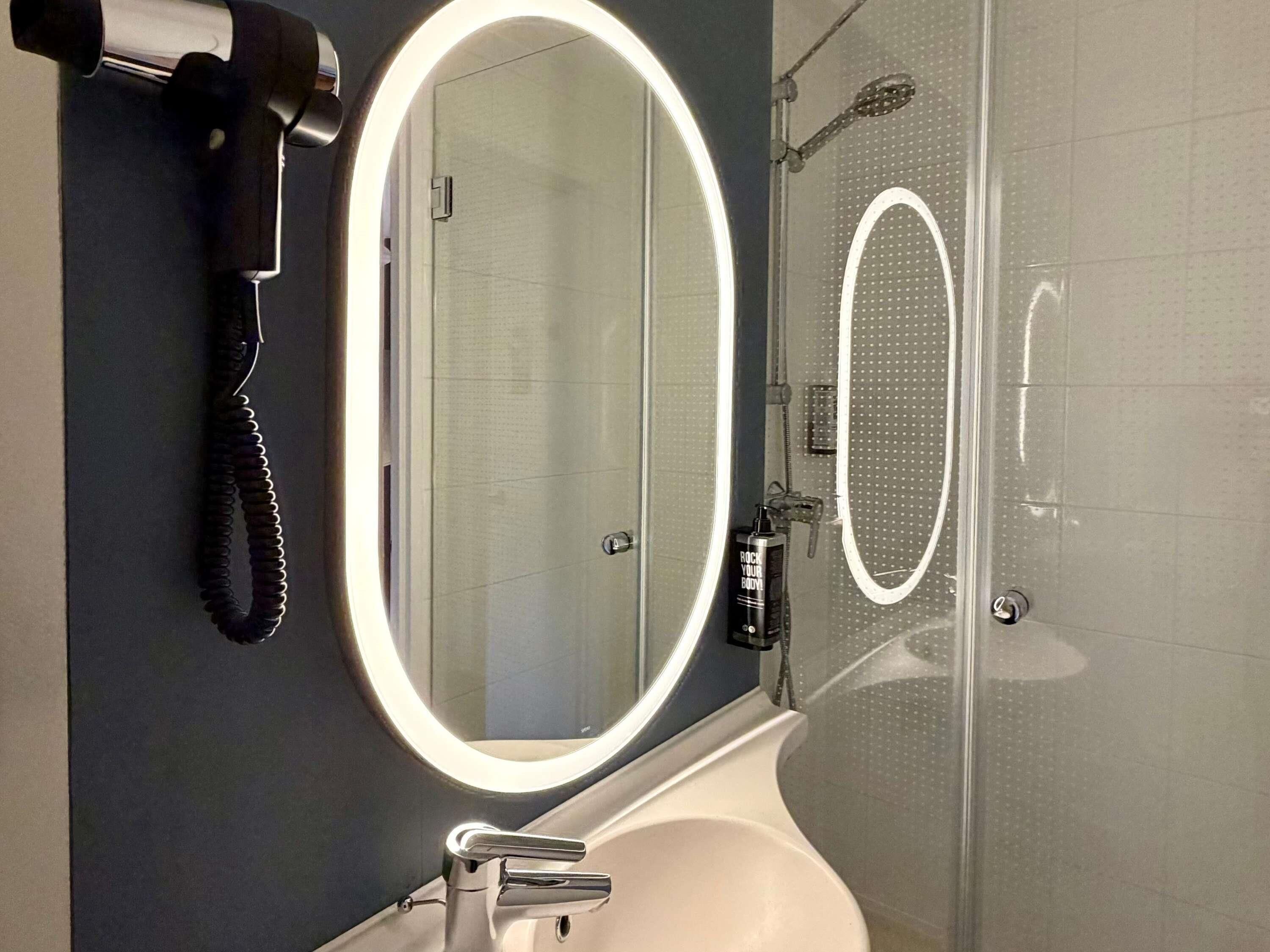 A bathroom with a large mirror, a handheld hair dryer, and a shower with a circular light.