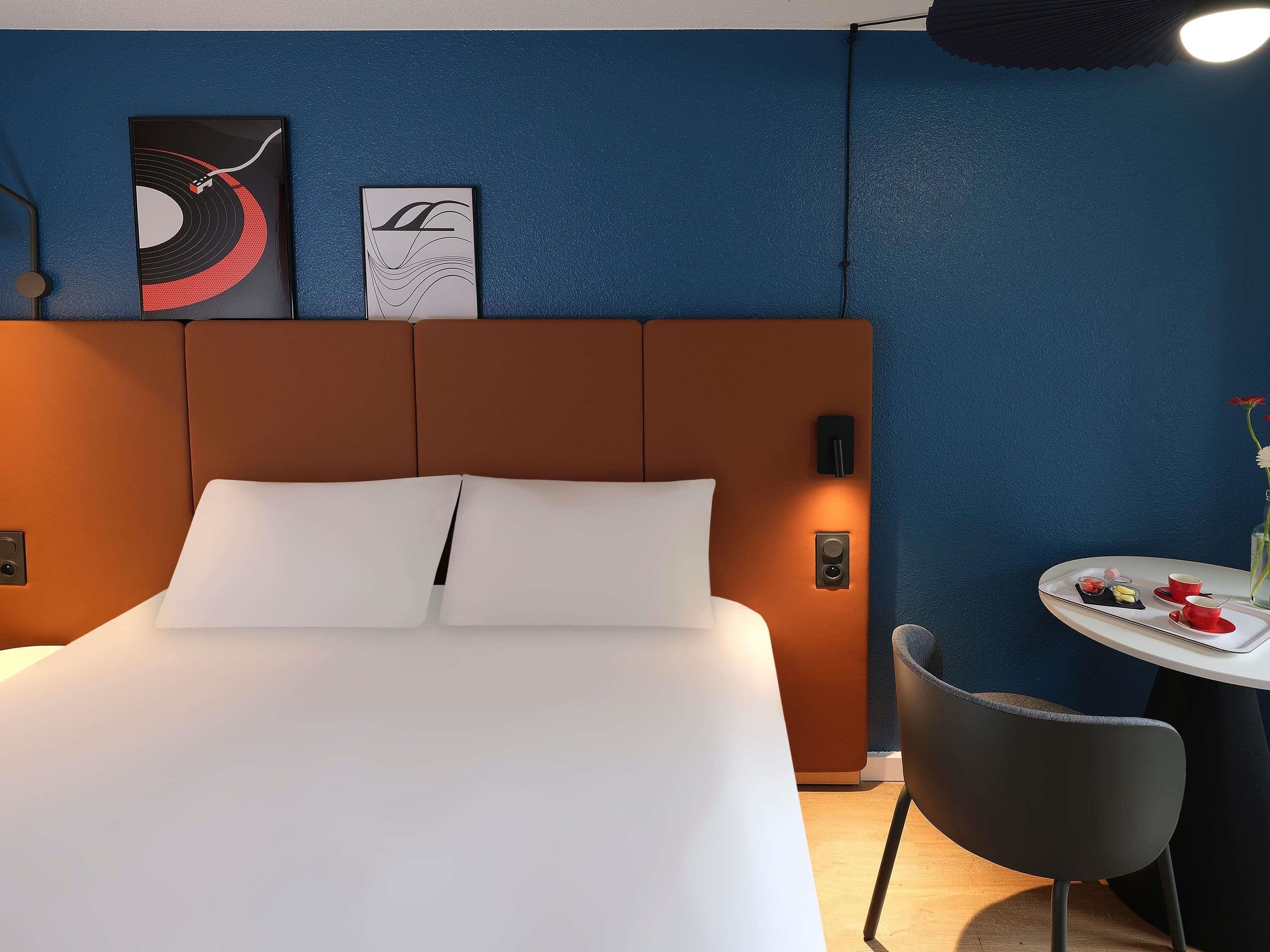 Una camera d'albergo con un letto, un comodino con un vassoio di cibo, una sedia e un'opera d'arte appesa al muro.