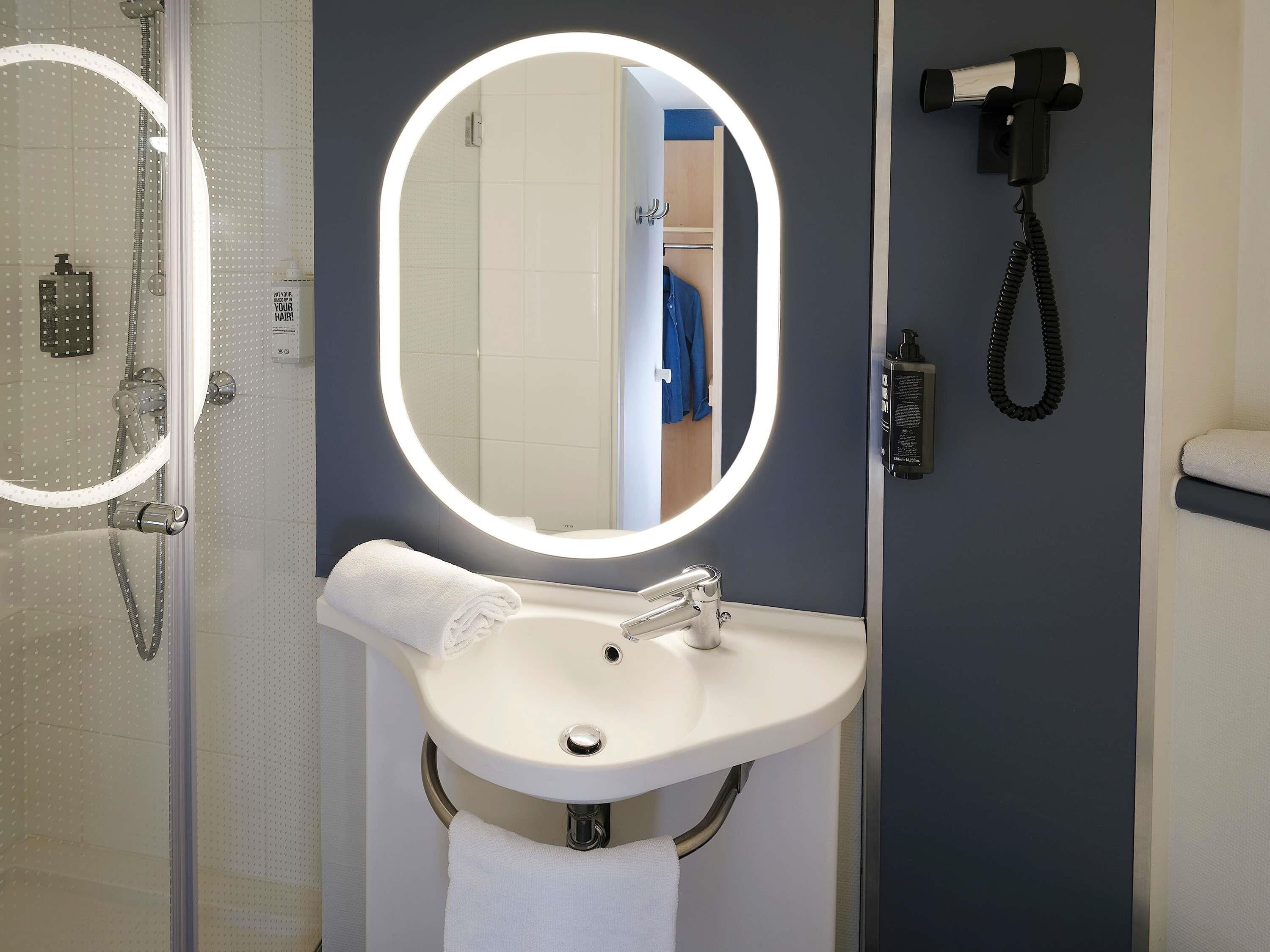 Un bagno con un lavandino bianco, uno specchio rotondo con luci, un soffione doccia a mano e un asciugacapelli fissato alla parete.