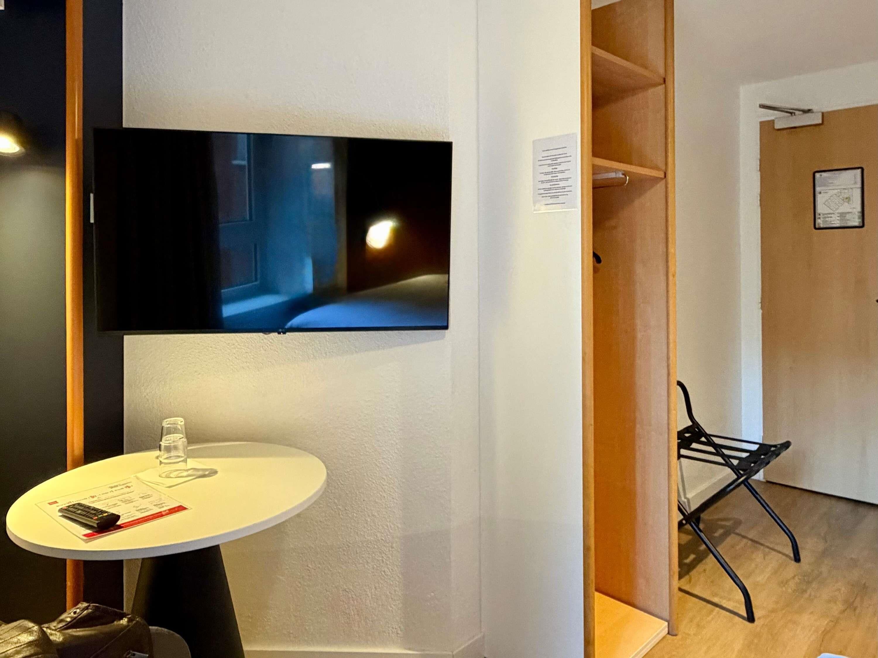 A room with a mounted TV, a small round table with a remote, a chair, and a wooden door.