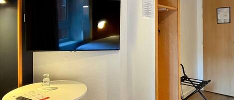 A room with a mounted TV, a small round table with a remote, a chair, and a wooden door.