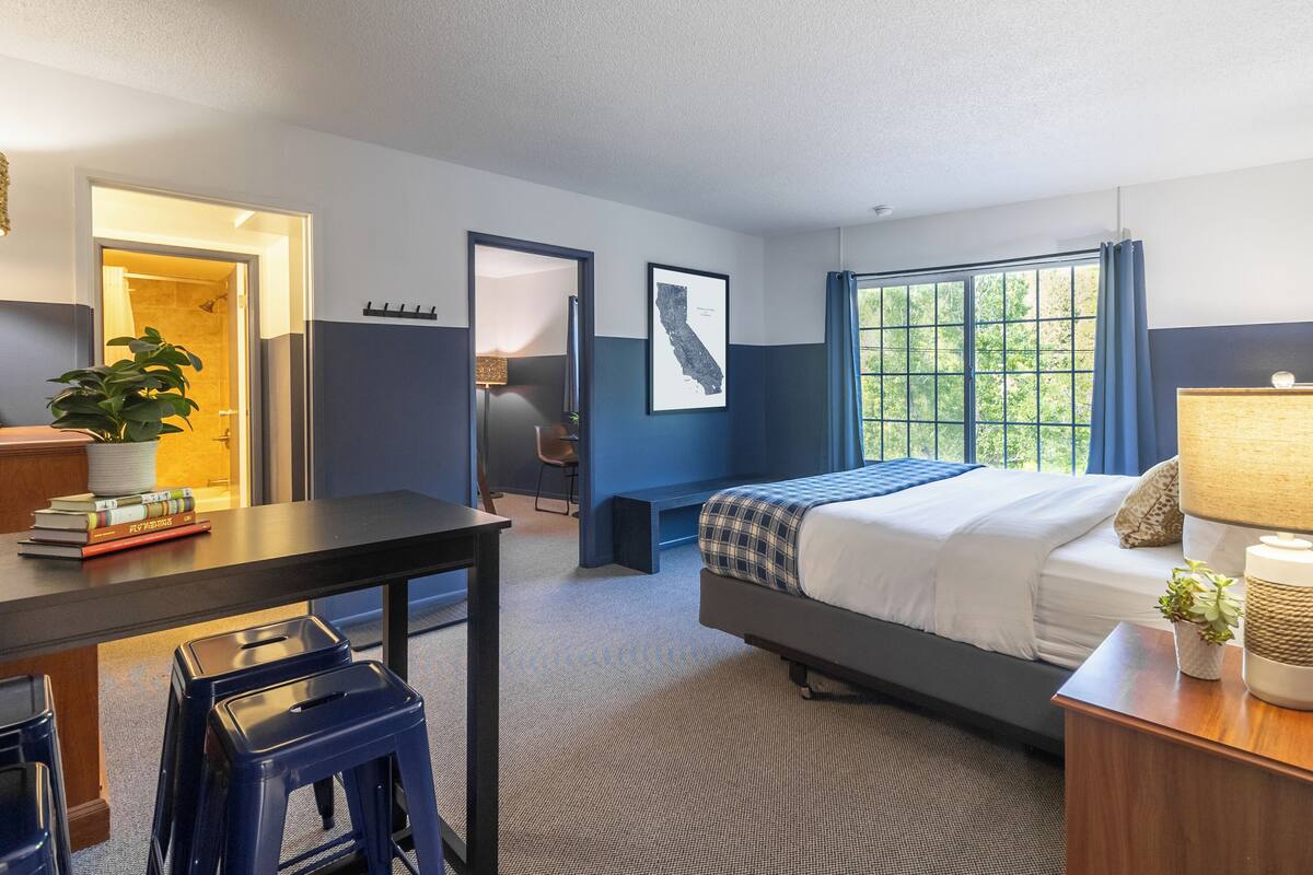 A hotel room with a bed, desk, chair, and a large window.